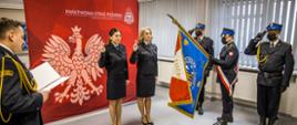 Str. Monika Wójcik- Janczak oraz str. Barbara Falarz składają ślubowanie na sztandar KW PSP w Opolu w obecności Opolskiego Komendanta Wojewódzkiego PSP w Opolu st. bryg. Krzysztofa Kędryka. W tle ślubujących plansza tła czerwonego z białym godłem Polski i białym napisem Państwowa Straż Pożarna Komenda Wojewódzka PSP w Opolu. Strażacy w umundurowaniu galowym oraz służbowym.