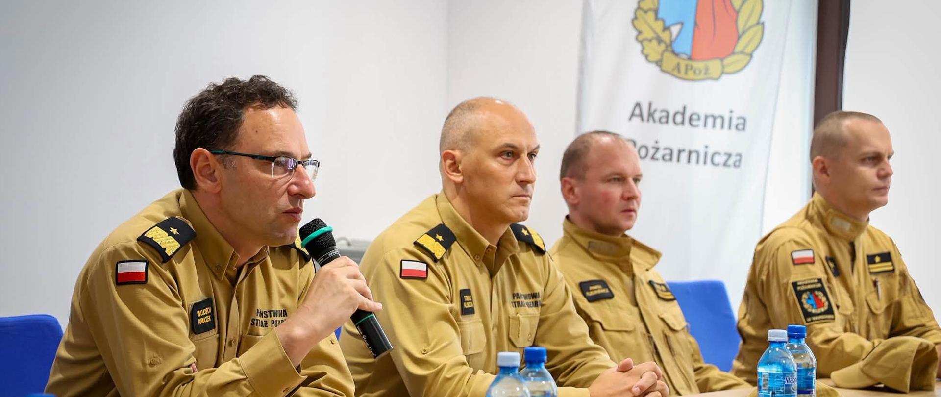 Czterech funkcjonariuszy Państwowej Straży Pożarnej siedzi przy stole podczas spotkania w Akademii Pożarniczej. Jeden z nich przemawia do mikrofonu.