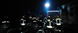 Noc. Strażacy idą w kierunku spalonego domu. Reflektor oświetla im drogę. Widać odblaskowe pasy na mundurach i hełmach.