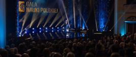 Zdjęcie od tyłu zaciemnionej sali, na podwyższeniu z przodu za mównicą stoi minister Kulasek i mówi do mikrofonu, za nim na granatowej ścianie wielki napis Gala Nauki Polskiej i reflektory.