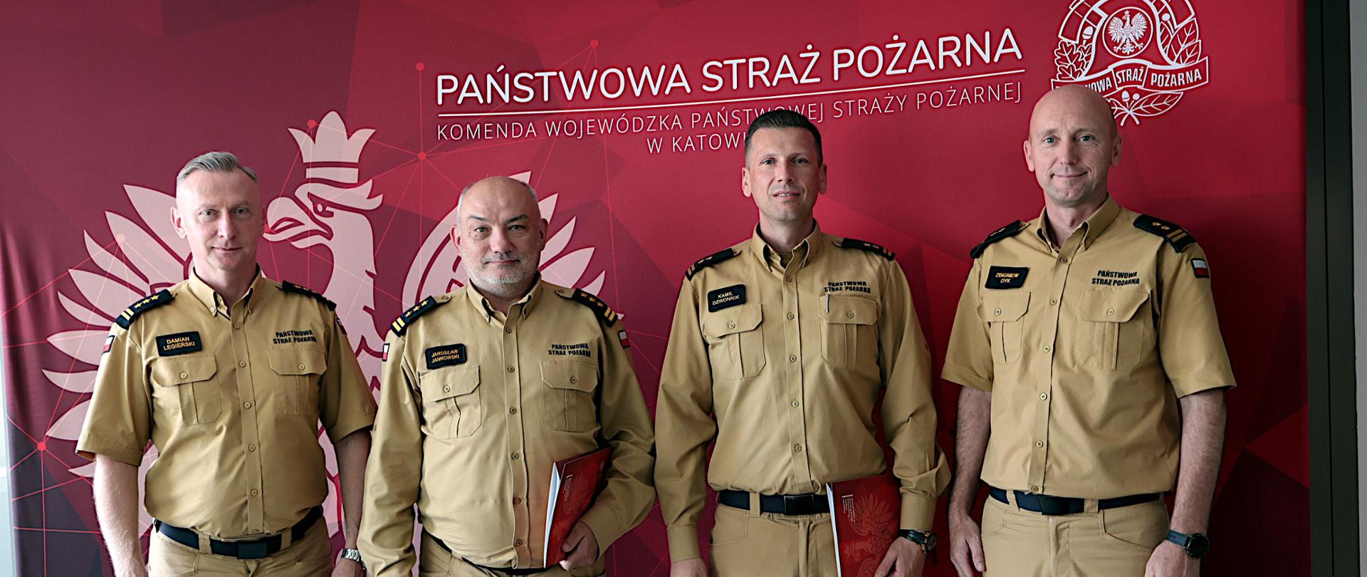 Na zdjęciu czterech funkcjonariuszy Państwowej Straży Pożarnej stoi obok siebie na tle czerwonej ścianki z dużym godłem i napisem Komendy Wojewódzkiej Państwowej Straży Pożarnej w Katowicach. Od lewej strony śląski komendant wojewódzki Państwowej Straży Pożarnej, st. bryg. Damiana Legierskiego, komendant Centralnej Szkoły Państwowej Straży Pożarnej w Częstochowie, st. bryg. Jarosław Jankowski, komendant miejski Państwowej Straży Pożarnej w Częstochowie, bryg. Kamil Dzwonnik oraz zastępca śląskiego komendanta bryg. Zbigniew Dyk. Wszyscy ubrani są w beżowe mundury służbowe i patrzą w stronę aparatu. Dwóch strażaków stojących pośrodku trzyma czerwone teczki z dokumentami.