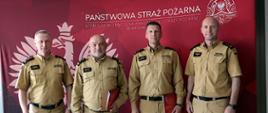 Na zdjęciu czterech funkcjonariuszy Państwowej Straży Pożarnej stoi obok siebie na tle czerwonej ścianki z dużym godłem i napisem Komendy Wojewódzkiej Państwowej Straży Pożarnej w Katowicach. Od lewej strony śląski komendant wojewódzki Państwowej Straży Pożarnej, st. bryg. Damiana Legierskiego, komendant Centralnej Szkoły Państwowej Straży Pożarnej w Częstochowie, st. bryg. Jarosław Jankowski, komendant miejski Państwowej Straży Pożarnej w Częstochowie, bryg. Kamil Dzwonnik oraz zastępca śląskiego komendanta bryg. Zbigniew Dyk. Wszyscy ubrani są w beżowe mundury służbowe i patrzą w stronę aparatu. Dwóch strażaków stojących pośrodku trzyma czerwone teczki z dokumentami.
