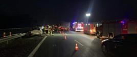 Zdjęcie przedstawia samochód stojący po lewej stronie jezdni po uderzeniu w barierkę. Obok stoją strażacy. Jeden pas ruchu oddzielony jest pachołkami. Po prawej stronie jadą inne samochody, a na poboczu stoją wozy strażackie. teren akcji oświetlony z jednego masztu. 