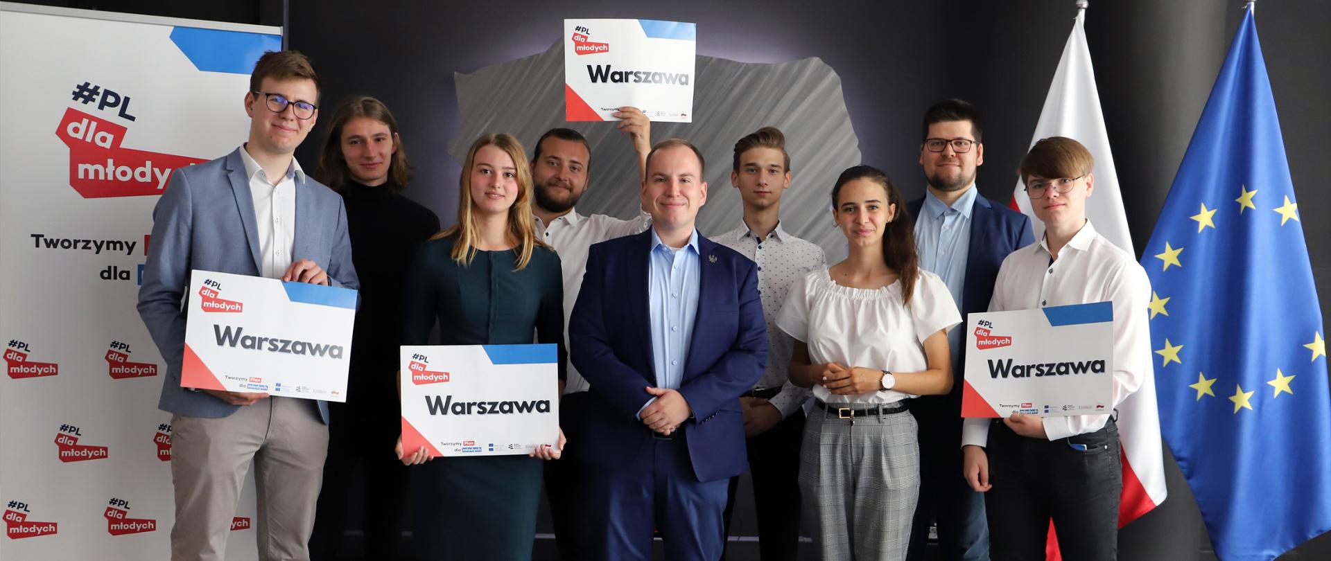 Wiceminister Adam Andruszkiewicz w otoczeniu przedstawicieli projektu #PLdlaMłodych (8 osób). Trzy osoby trzymają w dłoniach plansze z napisem Warszawa i logiem projektu. 