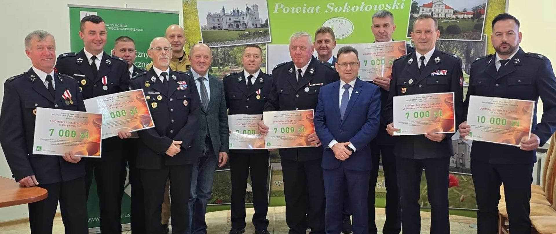 
Na zdjęciu znajduje się grupa trzynastu mężczyzn wewnątrz jasnego pomieszczenia, prawdopodobnie podczas uroczystości związanej z przyznawaniem funduszy dla straży pożarnej, na co wskazuje tło i rekwizyty.
Większość mężczyzn ubrana jest w mundury galowe straży pożarnej w kolorze ciemnym (granatowym lub czarnym) z emblematami i medalami. Dwóch mężczyzn, stojących w centrum, jest ubranych w garnitury (jeden szary, drugi granatowy). Wszyscy stoją twarzą do aparatu.
Pomiędzy mężczyznami widać kartony lub symboliczne czeki z napisami w języku polskim, wskazującymi na kwoty pieniężne i przeznaczenie na 'OSP' (Ochotnicza Straż Pożarna) lub 'PSP' (Państwowa Straż Pożarna), np. "DOTACJA / ŚRODKI DLA / OCHOTNICZEJ STRAŻY POŻARNEJ" z kwotami takimi jak 7000 zł i 10000 zł. Mężczyźni trzymają te czeki w rękach.
W tle, za mężczyznami, widać baner reklamowy lub plakat z napisem "Powiat Sokołowski" oraz grafiką przedstawiającą zamek lub pałac, co sugeruje lokalizację wydarzenia. Podłoga jest jasna, a z prawej strony widać fragmenty krzeseł. Oświetlenie jest jasne.
Mężczyźni mają poważne lub lekko uśmiechnięte miny i sprawiają wrażenie uczestniczących w oficjalnym wydarzeniu.