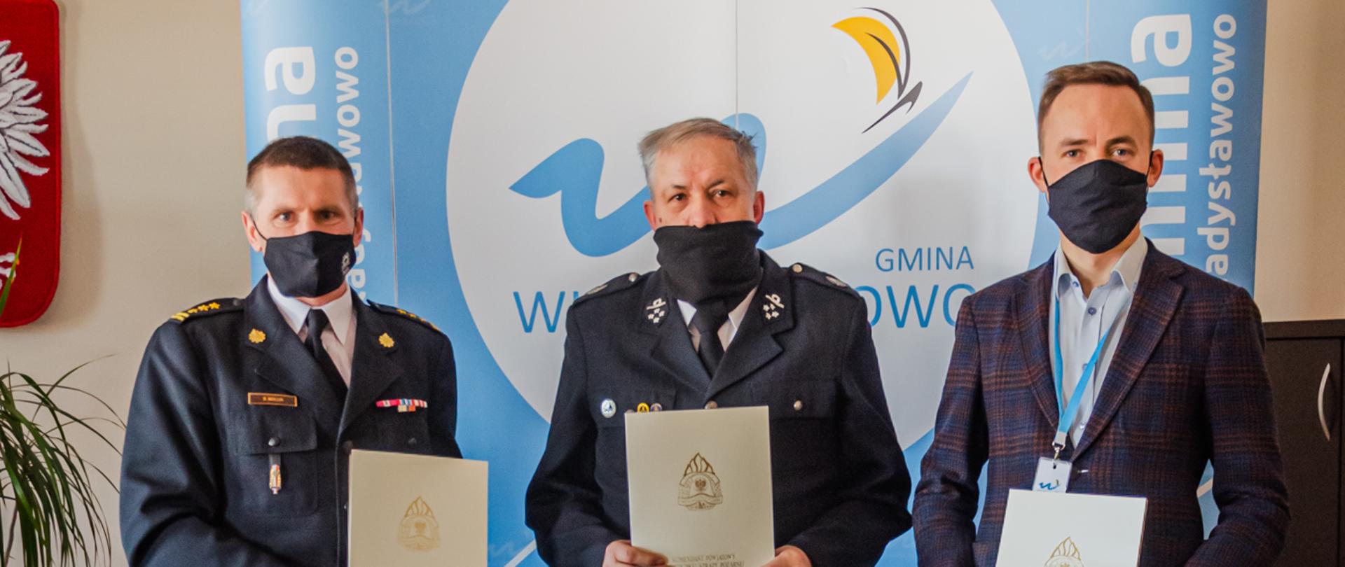 Zdjęcie przedstawia trzech stojących mężczyzn z tego dwóch w mundurach od lewej strażak państwowej straży pożarnej i druh ochotniczej straży pożarnej, a po prawej stoi młody mężczyzna w marynarce - burmistrz Władysławowa. Mężczyźni trzymają przed sobą ozdobne kartki.