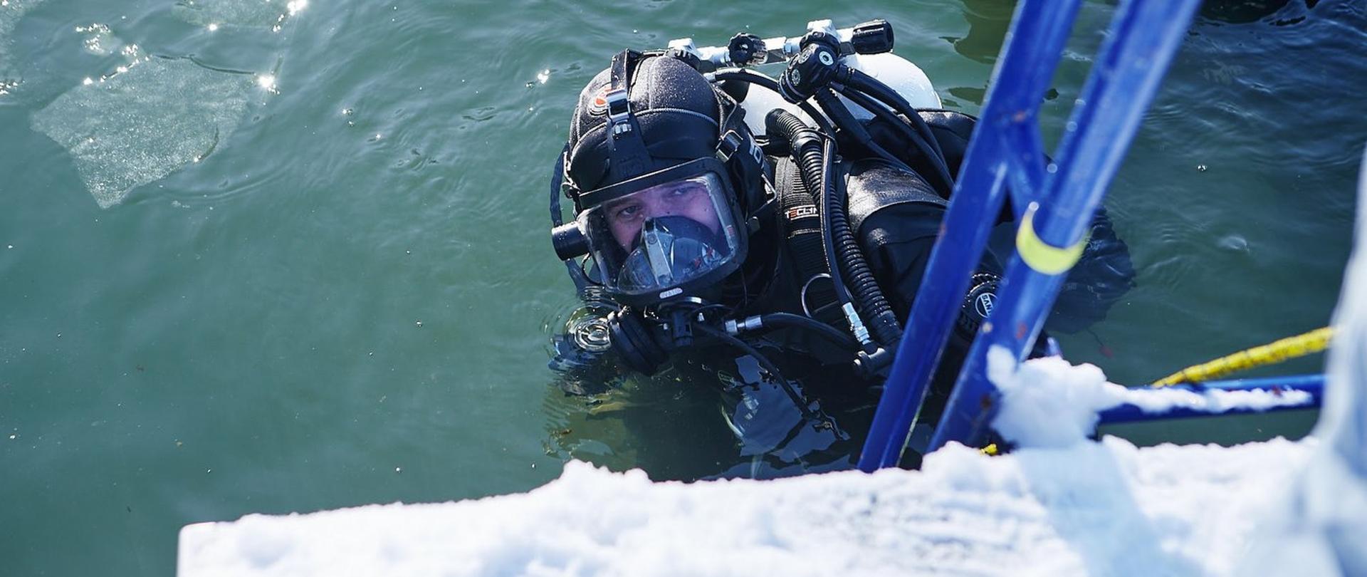 Zdjęcie zrobione w zimie, na zewnątrz przy zbiorniku wodnym. Płetwonurek zanurzony jest w wodzie w której pływają tafle lodu. 