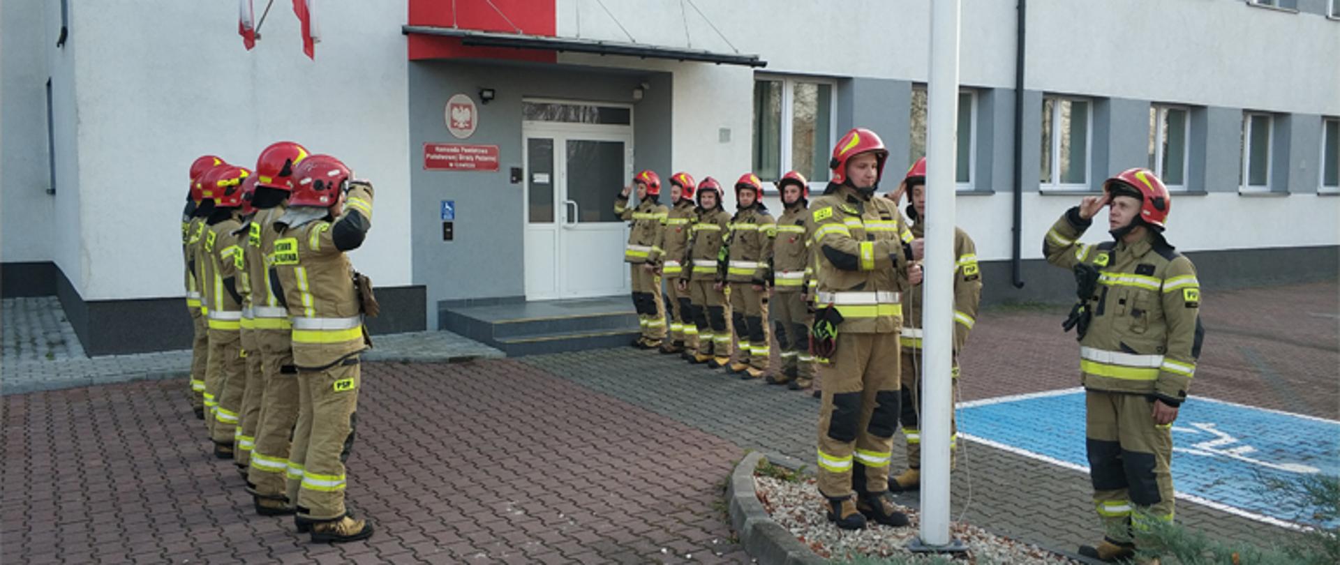 szesnastu strażaków ubranych w umundurowanie bojowe barwy żółto-brązowej z hełmem koloru czerwonego uczestniczy w uroczystym podniesieniu flagi państwowej na maszt, strażacy stoją naprzeciw siebie po lewej stronie siedem osób a po prawej sześć osób, przy maszcie flagowym stoją trzy osoby dwie salutują a jedna wciąga flagę na szczyt masztu, w tle budynek komendy powiatowej państwowej straży pożarnej w Łowiczu