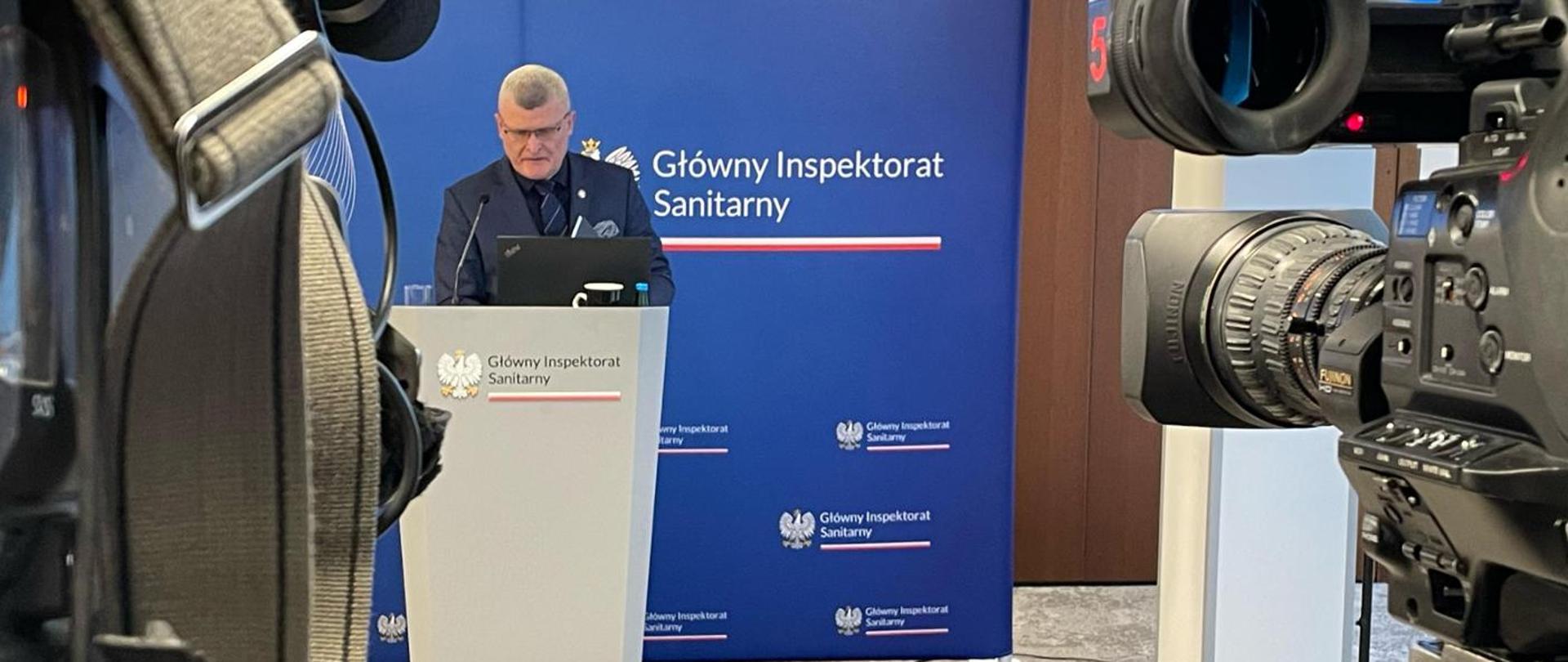 W centralnej części zdjęcia Główny Inspektor Sanitarny stojący przy mównicy, w tle niebieski plakat z napisem "Główny Inspektorat Sanitarny". Po prawej części zdjęcia kamera skierowana w stronę Głównego Inspektora Sanitarnego.
