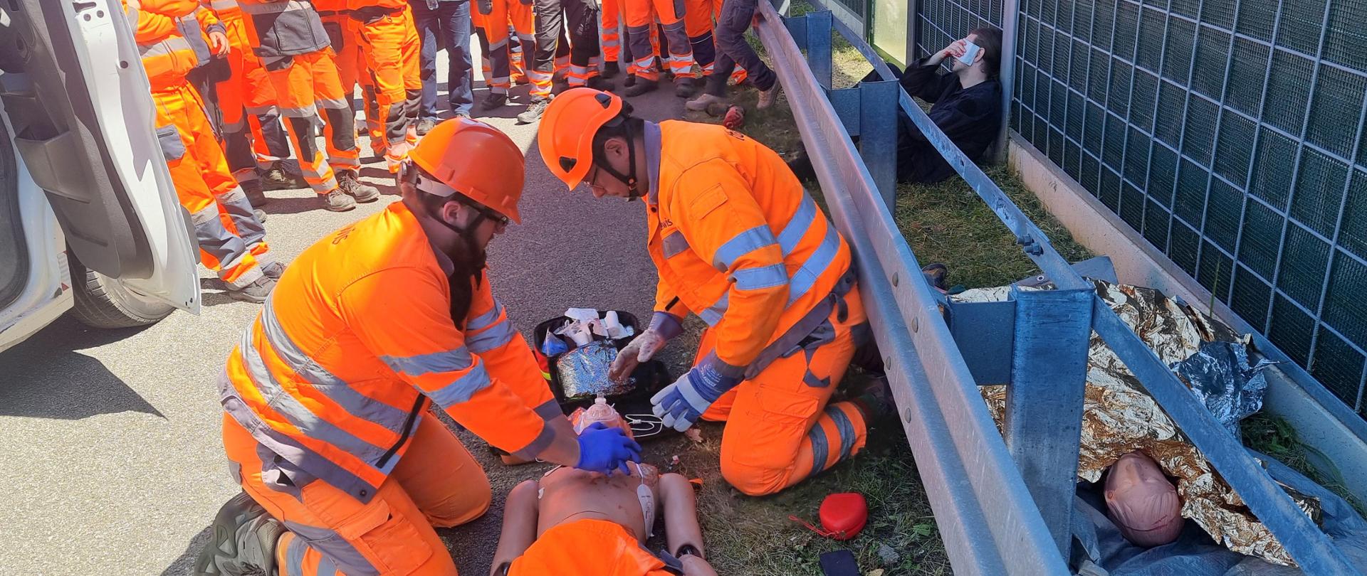 Szkolenie z pierwszej pomocy - dwóch drogowców w pomarańczowych ubraniach ochronnych i kaskach wykonuje na fantomie masaż serca i sztuczne oddychanie