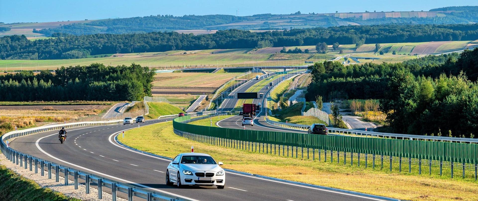 Droga ekspresowa S7 na odcinku Moczydło - Miechów. Dwujezdniowa droga ekspresowa wijąca się między polami i lasami po niewielkich pagórkach. Widoczne pojazdy poruszające się w obu kierunkach.