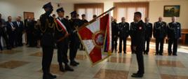 Nowy Komendant Powiatowy PSP w Skarżysku-Kamiennej wita sie ze sztandarem komendy. Poczet salutuje. W tle widzimy strażaków na zbiórce. Wydarzenie ma miejsce w pomieszczeniu.