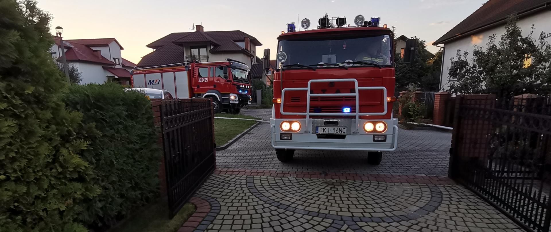 Zdjęcie przedstawia dwa samochody pożarnicze, które stoją przy wjeździe na prywatną posesję gdzie wybuchł pożar.