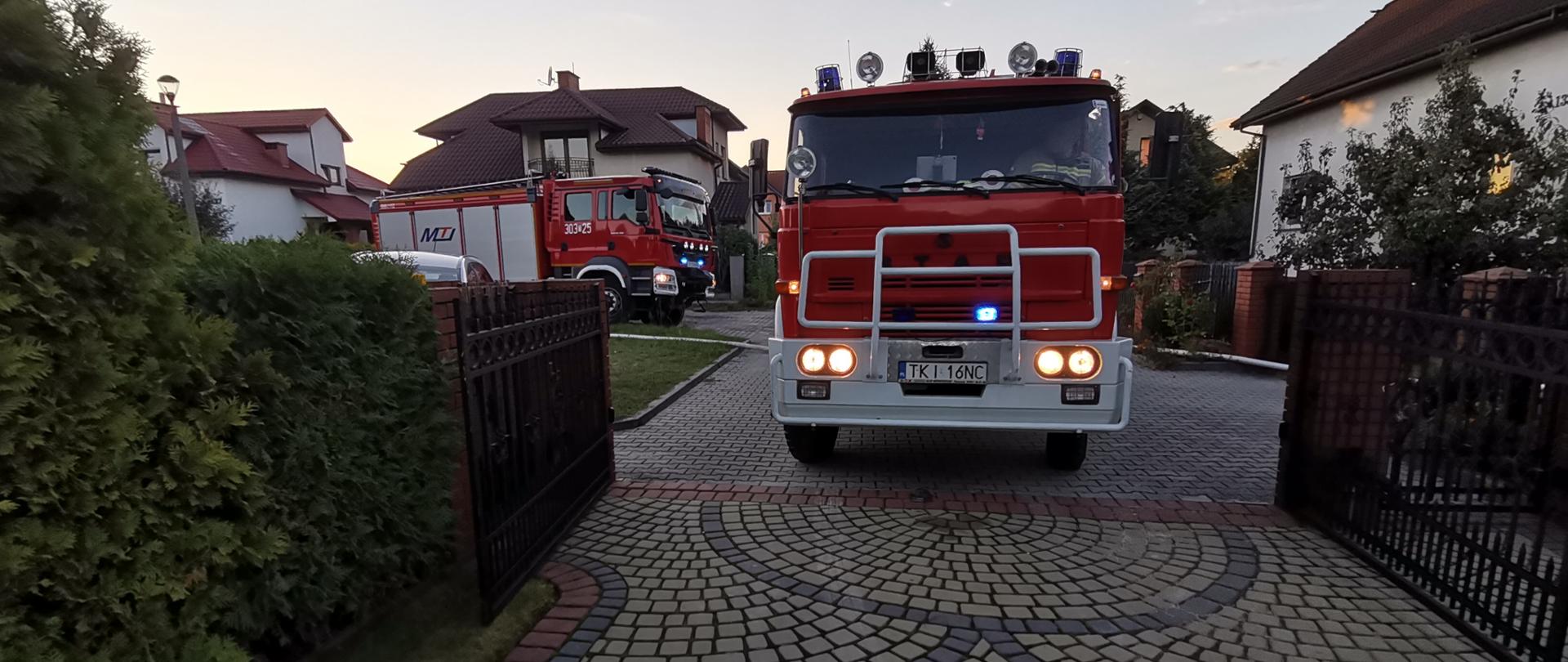 Zdjęcie przedstawia dwa samochody pożarnicze, które stoją przy wjeździe na prywatną posesję gdzie wybuchł pożar.