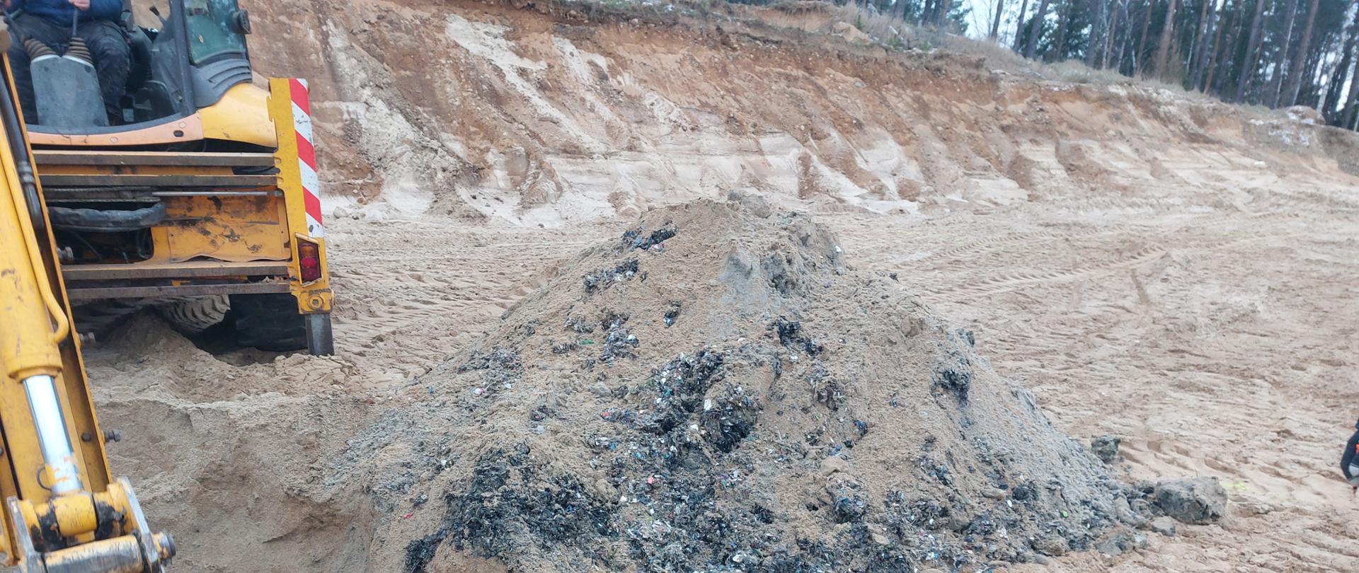Odkopane odpady lezą na piasku, obok koparka