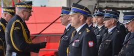 Zastępca komendanta głównego PSP wraz z komendantem wojewódzkim PSP wręczają odznaczenia/awanse stojącym przed nimi strażakom