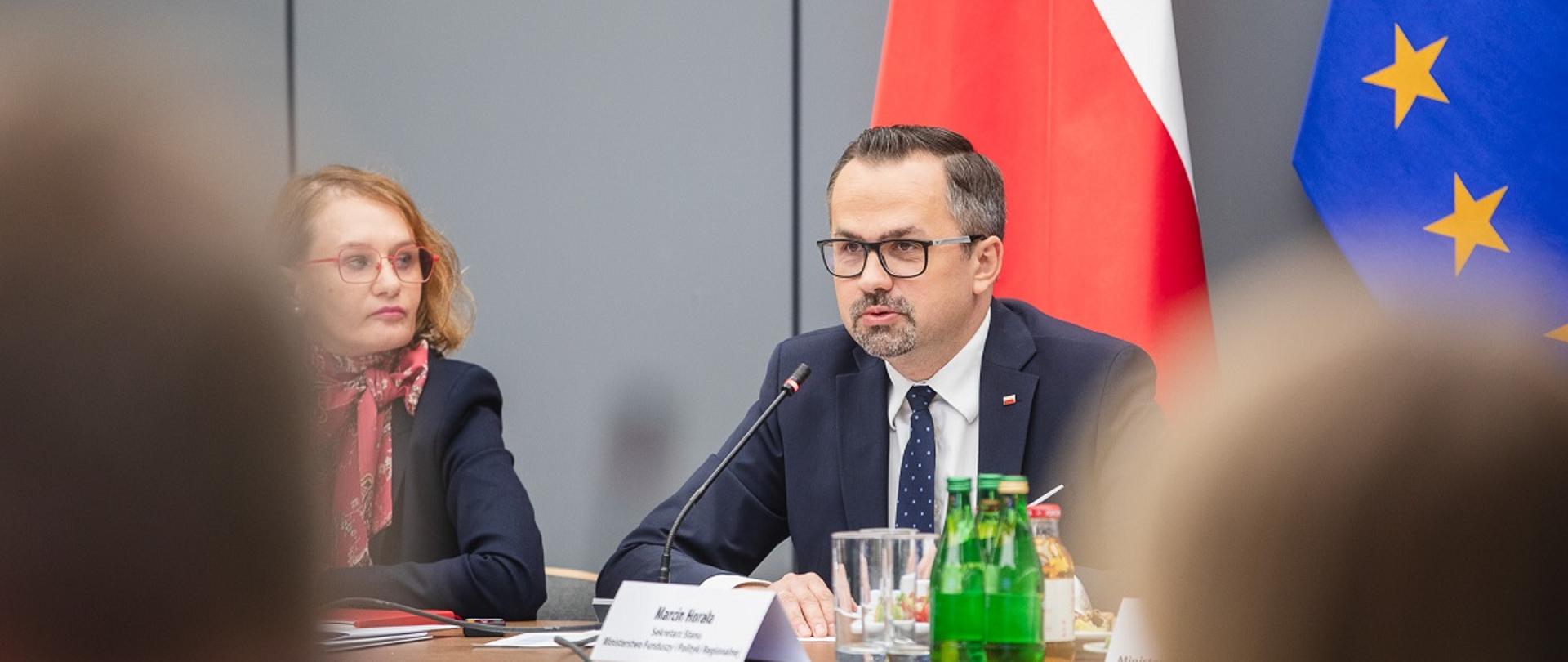 Pierwsze posiedzenie Komitetu Monitorującego Krajowy Plan Odbudowy - wiceminister funduszy i polityki regionalnej Marcin Horała siedzi przy stole i przemawia przez mikrofon, po jego lewej stronie siedzi dyrektor Departamentu Strategii w MFiPR Renata Calak. 