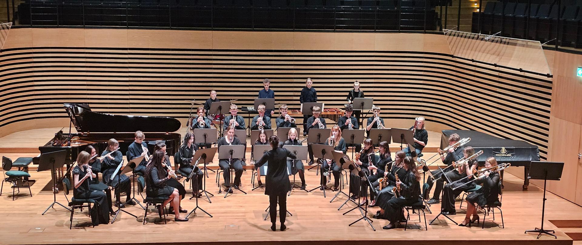 Trzydzieścioro uczniów ubranych na czarno siedzi w półkolu w trzech rzędach i grają na instrumentach dętych i perkusyjnych, w środku tyłem do widowni stoi kobieta i dyryguje. Po prawej i lewej stronie za orkiestrą stoją dwa fortepiany.