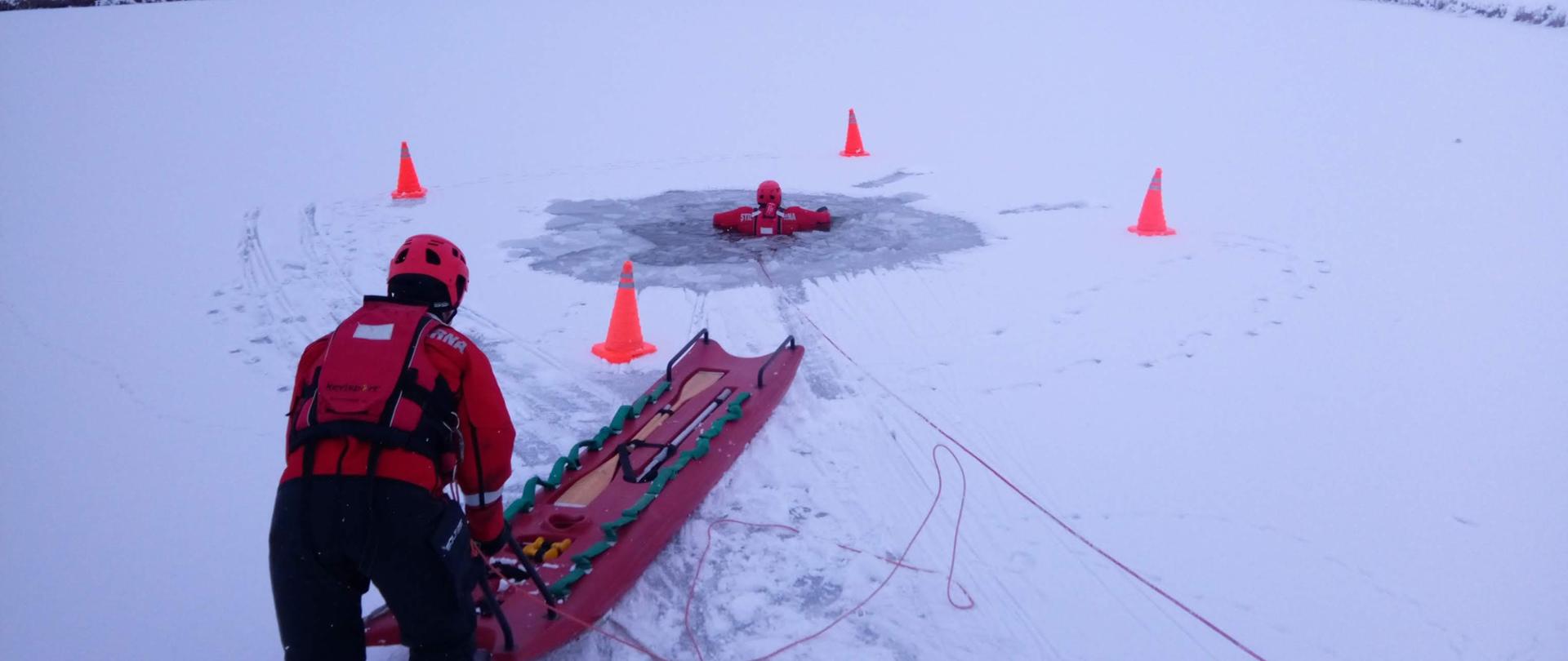 Podejmowanie osoby topiącej, która znajduje się w przerębli zamarzniętego zbiornika przez strażaka zabezpieczonego linką ratowniczą, ubranego w ubranie do pracy w wodzie przy użyciu sań lodowych , w tle zabudowania miejscowości.