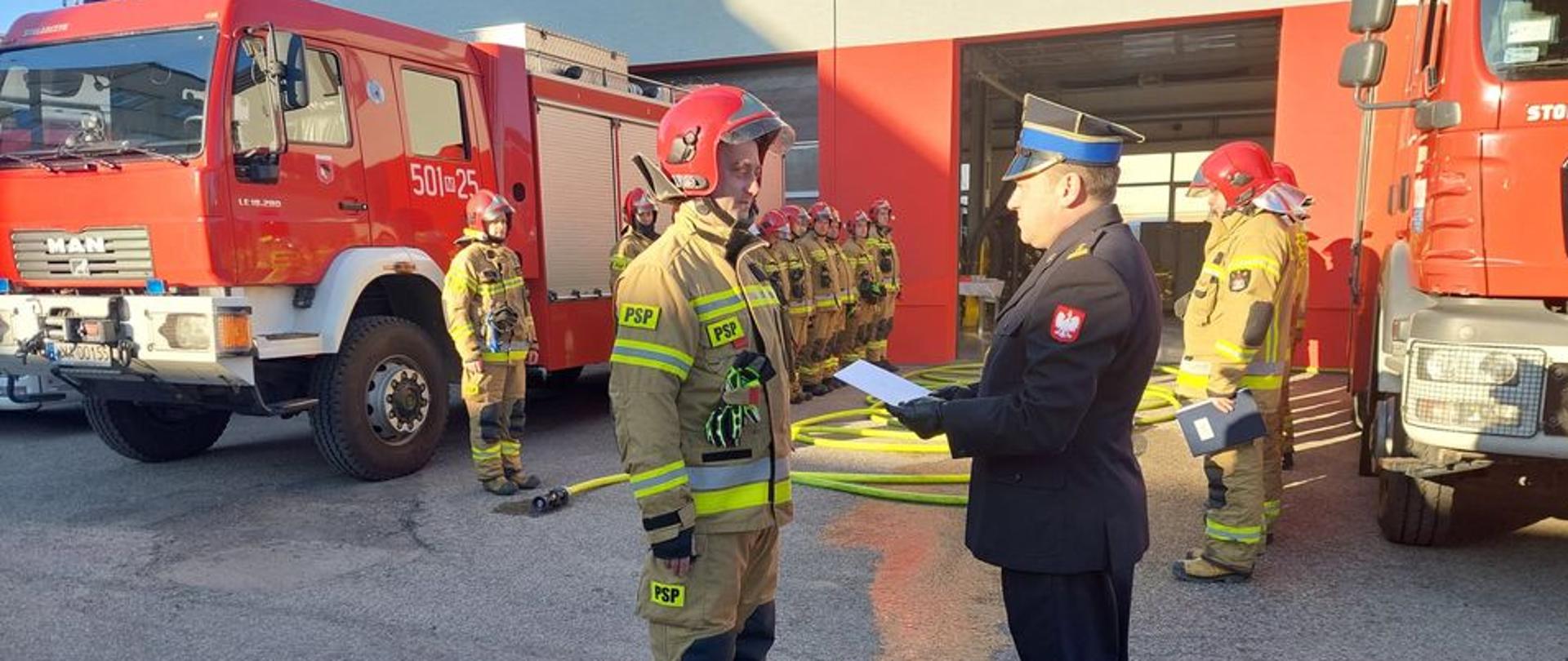 Komendant Powiatowy wręcza teczkę funkcjonariuszowi. W tle zmiany służbowe stoją naprzeciwko siebie.