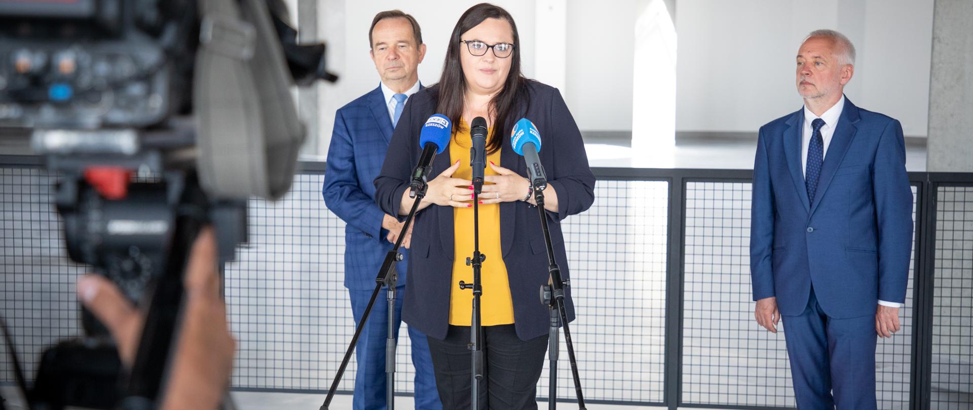 Przed mikrofonem stoi wiceminister Małgorzata Jarosińska-Jedynak. Za nią dwóch mężczyzn. Przed nimi kamera.