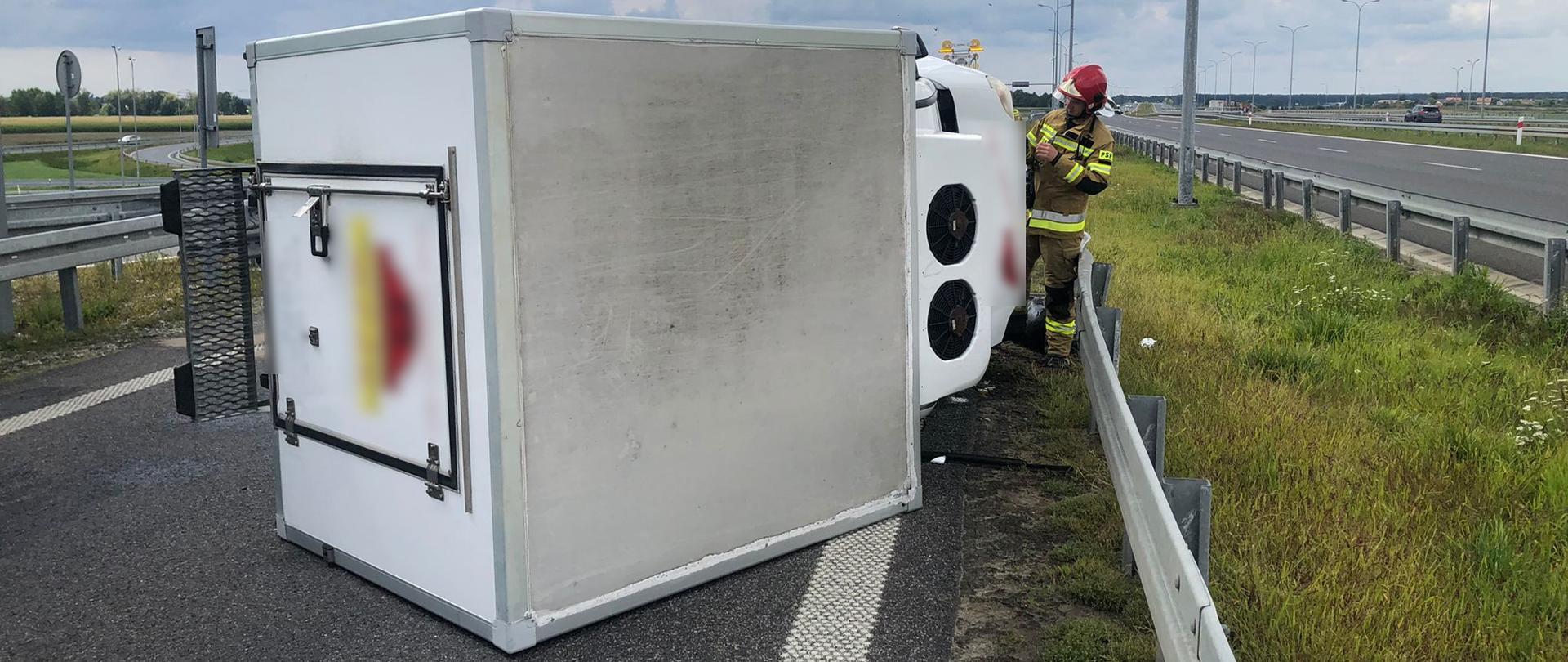 widoczne miejsce zdarzenia, biały samochód dostawczy leży na pasie ruchu trasy S8. Widoczny strażak prowadzący działania.