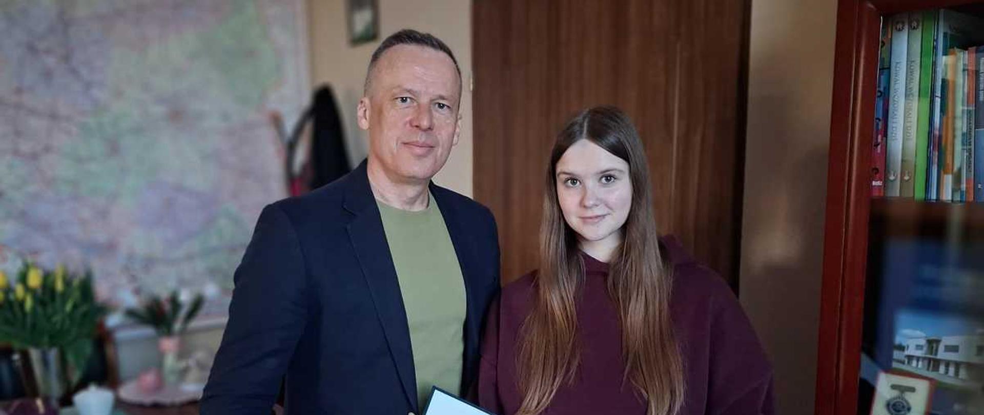 Zdjęcie przedstawia stojących obok siebie dorosłego mężczyznę i młodą kobietę w pomieszczeniu, prawdopodobnie podczas ceremonii wręczenia dyplomu. Oboje patrzą prosto w obiektyw.
Mężczyzna: Znajduje się po lewej stronie. Jest ubrany w ciemną marynarkę nałożoną na zieloną koszulkę. Ma krótkie, siwiejące włosy i lekki uśmiech.
Kobieta: Znajduje się po prawej stronie. Jest młodą osobą z długimi, brązowymi włosami. Ma na sobie bordową bluzę z kapturem. Również lekko się uśmiecha.
Dyplom: Oboje trzymają otwarty, oprawiony w czarną ramkę dokument. Widoczny tekst na dyplomie to ""DYPLOM"" oraz ""Nikoletty Gorczyńskiej"".
Tło: W tle, na ścianie za nimi, wisi duża mapa. Po prawej stronie widoczny jest regał z książkami i segregatorami. Po lewej stronie, na stole, znajdują się żółte tulipany w wazonie.