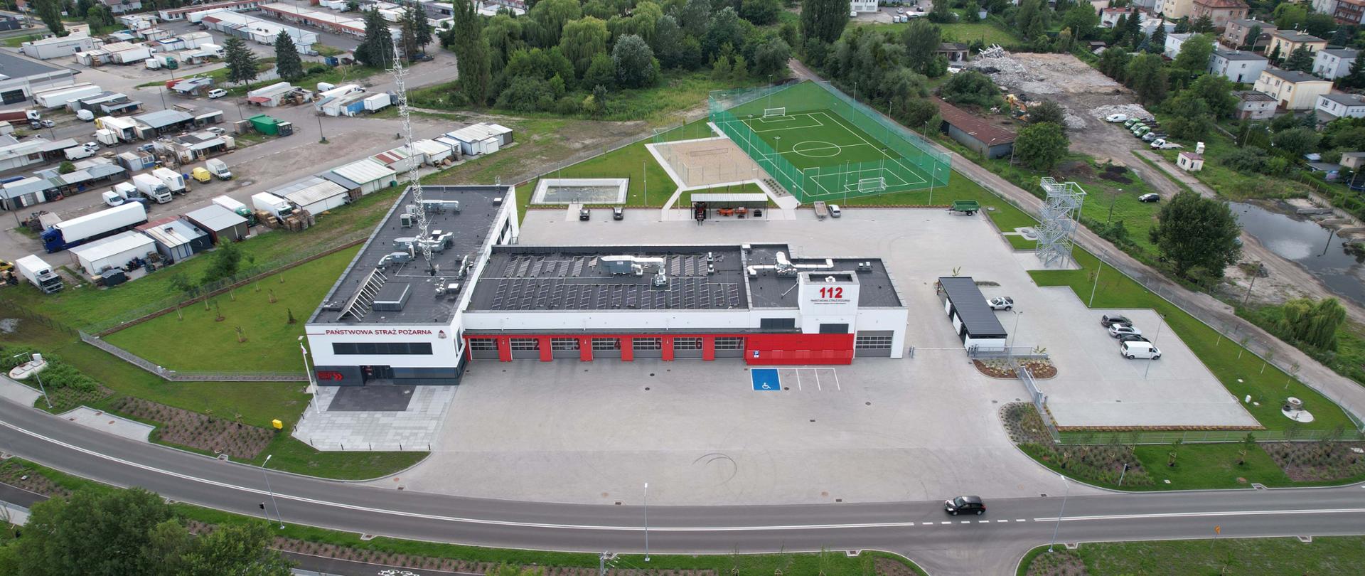 Szaro – czerwony budynek komendy i jednostki strażackiej z siwymi bramami wyjazdowymi oraz placami i boiskami sportowymi widziany od frontu z wysokości.