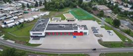 Szaro – czerwony budynek komendy i jednostki strażackiej z siwymi bramami wyjazdowymi oraz placami i boiskami sportowymi widziany od frontu z wysokości.
