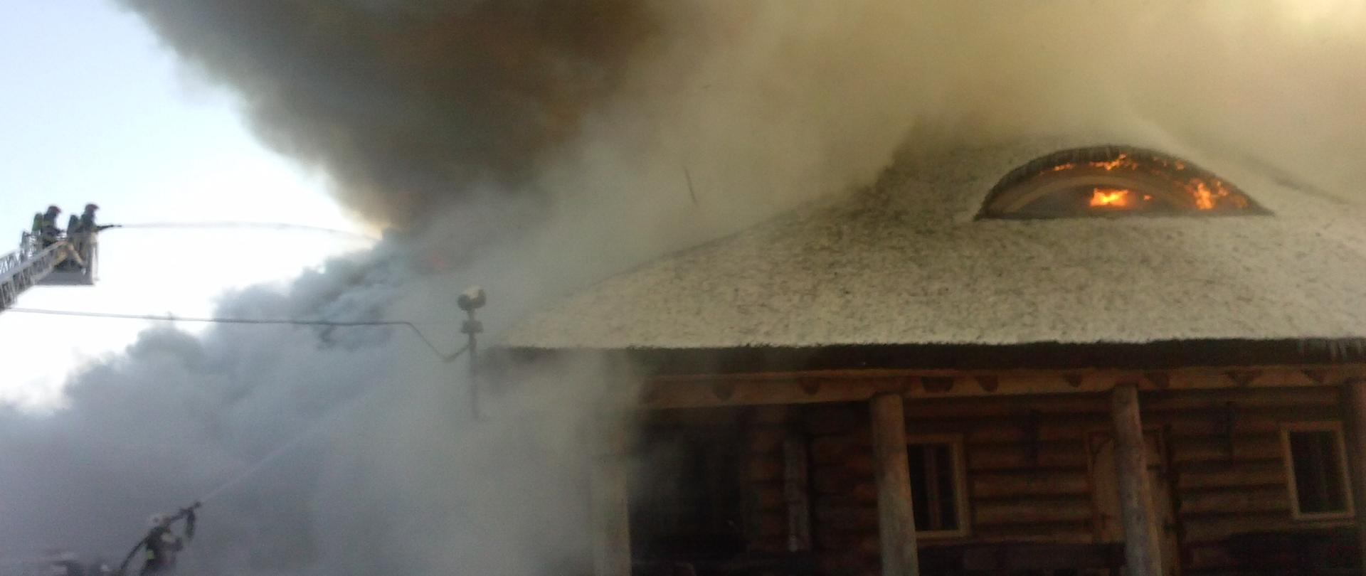 Budynek zbudowany z belek i siana płonie, strażacy gaszą pożar przy użyciu drabiny