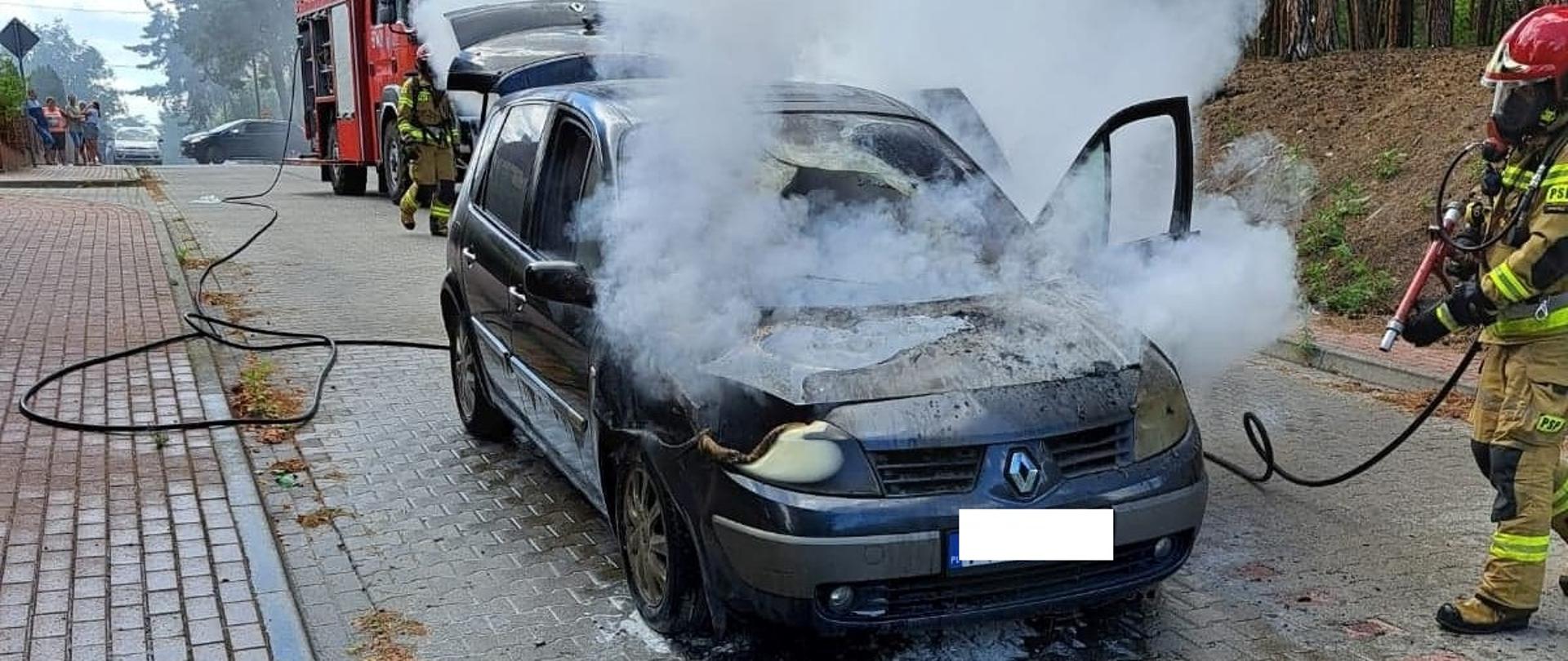 Pożar samochodu osobowego. Ulica Englerta w Rawiczu. Na jezdni stoi samochód osobowy koloru ciemnego spowity białym dymem. W tle fragment pojazdu straży pożarnej. Z przodu płonącego auta znajduje się strażak zabezpieczony sprzętem ochrony układu oddechowego, który gasi pożar. Auto posiada uszkodzoną szybę czołową i przednią część. Z lewej strony, w oddali na chodniku stoi grupka ludzi. Z prawej strony - lasek miejski. Pomiędzy palącym się samochodem a wozem strażackim znajduje się inny strażak.
