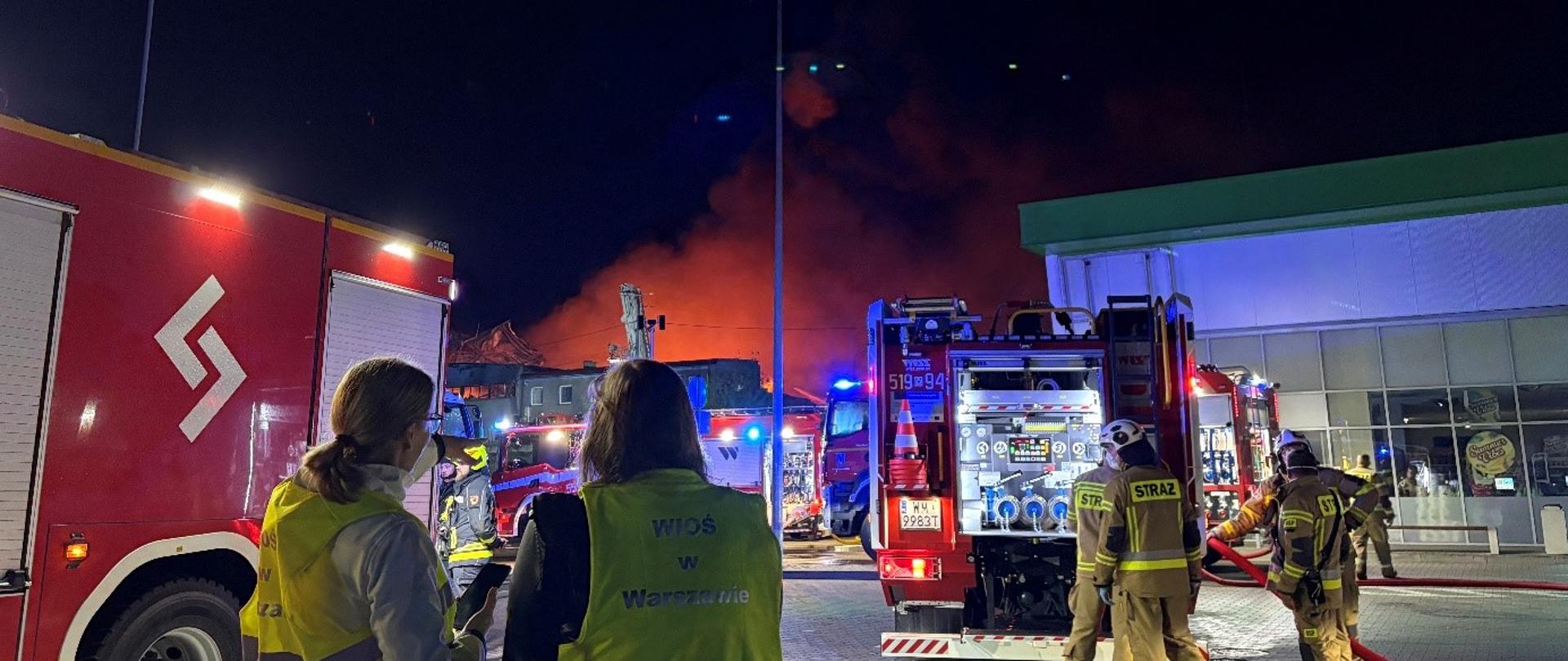 Dwie inspektorki Wojewódzkiego Inspektoratu Ochrony Środowiska w Warszawie prowadzą nocne działania na terenie pożaru. Obserwują kilku funkcjonariuszy Straży Pożarnej przy wozie bojowym. W tle widać parterowe zabudowania oraz czerwoną łunę pożaru na tle czarnego nieba.
