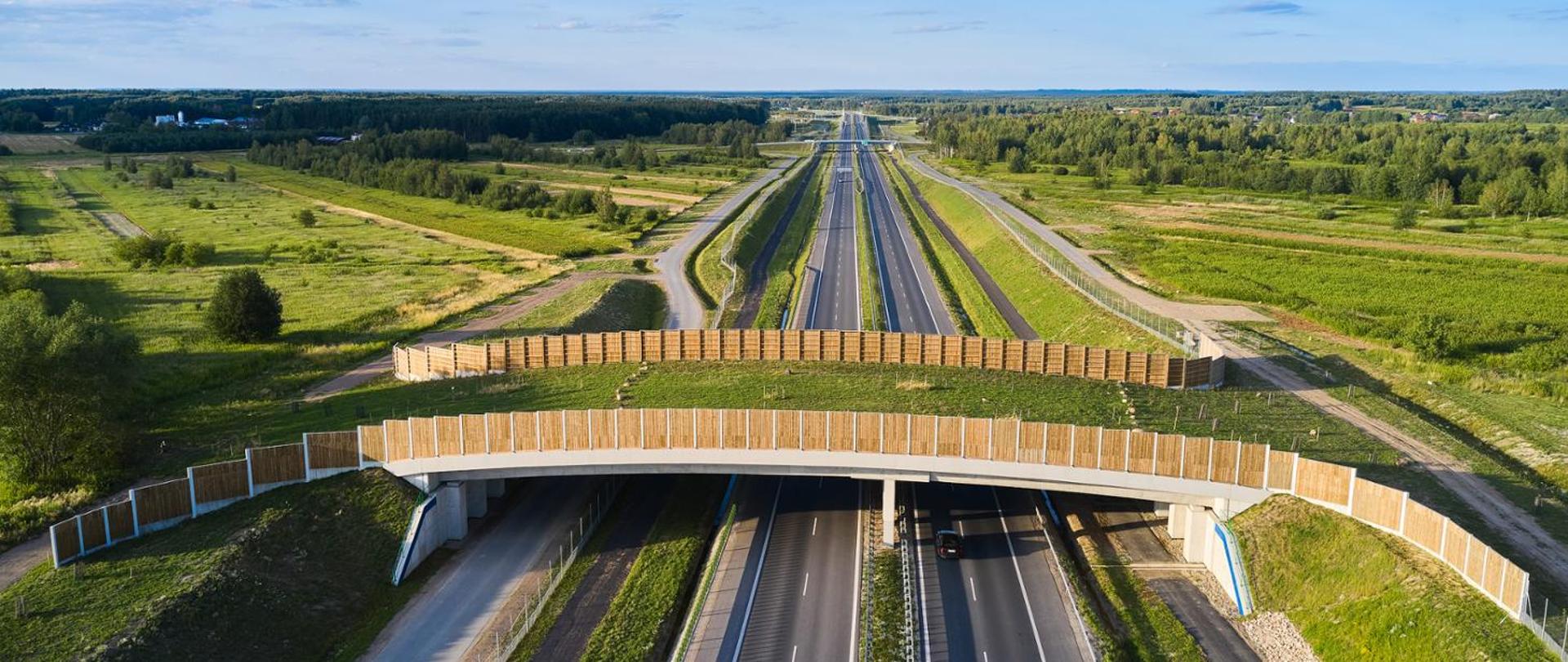 Widok z góry na drogę ekspresową. Na pierwszym planie widać przejście górne dla zwierząt. Dookoła lasy, świeci słońce. 