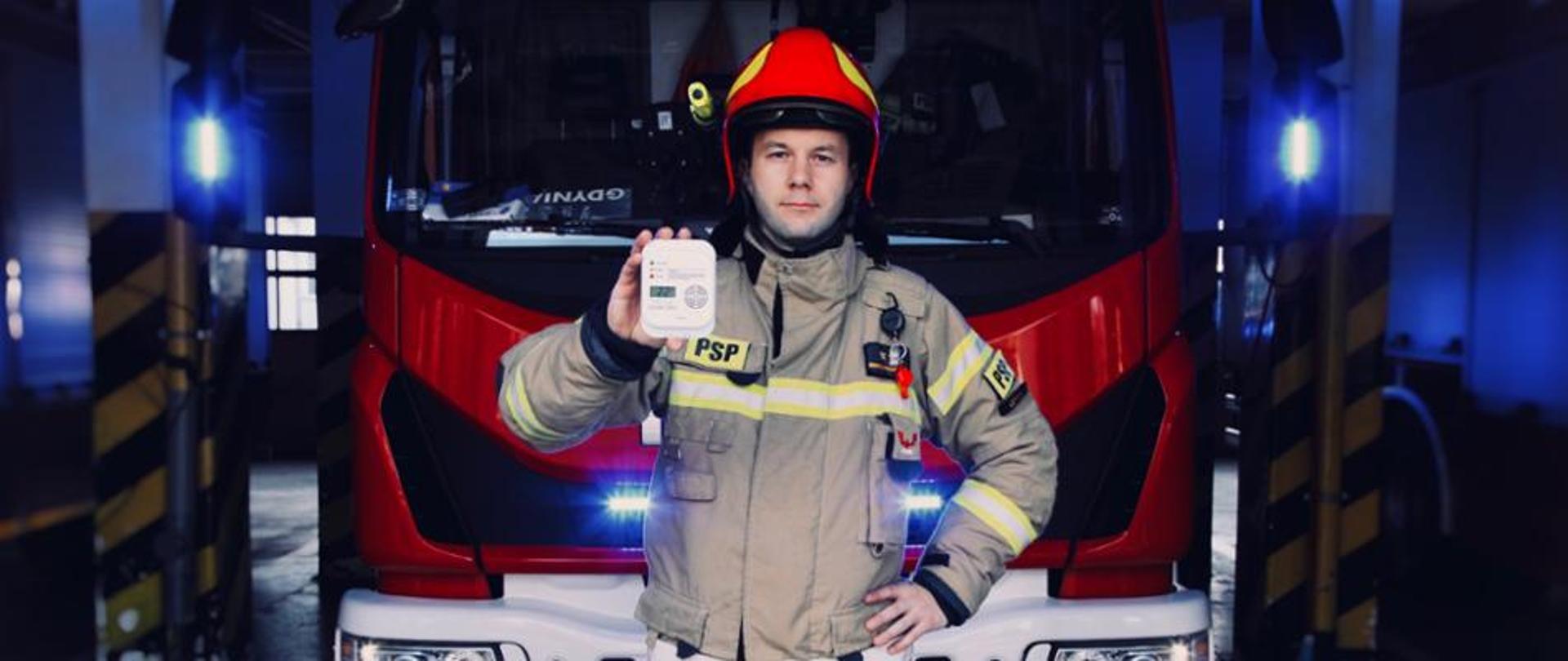 Zdjęcie przedstawia strażaka stojącego na tle samochodu pożarniczego, który trzyma w ręku czujkę. 