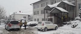 Kolorowe zdjęcie przedstawia na pierwszym planie, po lewej, tył srebrnego samochodu pożarniczego Państwowej Straży Pożarnej z napisem Straż na czerwonym pasem. Obok pojazdu stoją dwaj umundurowani strażacy. Na środkowym planie widoczne mocno zaśnieżone pojazdy osobowe na parkingu przy budynku ośrodka zdrowia. Po prawej stronie dwaj strażacy prowadzą starszą osobę, po stromych schodach do budynku. Na zdjęciu widoczna duża ilość śniegu.