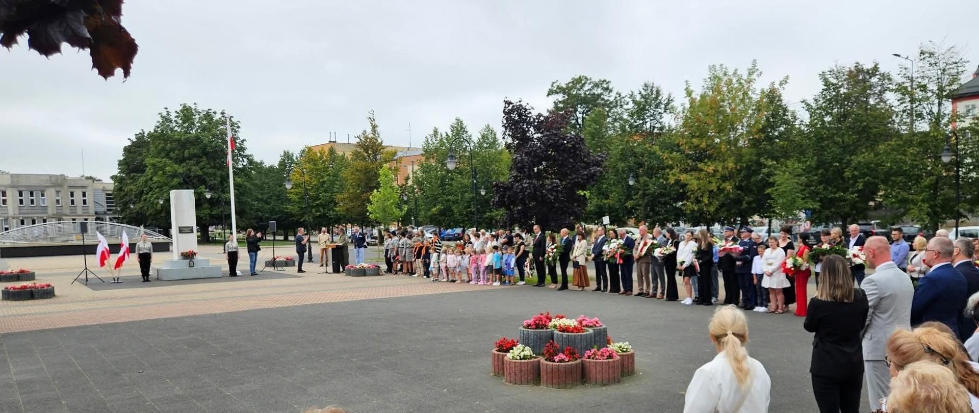 Obraz przedstawia grupę ludzi zebranych na placu miejskim rocznicę II wojny światowej. W tle widać pomnik i maszt z polską flagą. Niektóre osoby trzymają kwiaty, a kilka ma na sobie mundury. Wokół placu rosną drzewa, a w pobliżu stoją donice z kwiatami. 