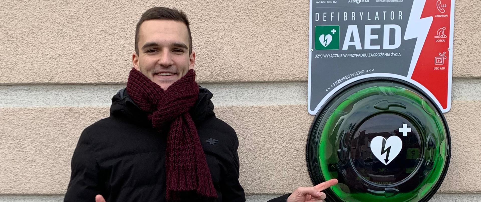 Po lewej stronie stoi pomysłodawca projektu AED i wskazuje na kapsułę, w której zamontowany jest defibrylator.