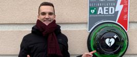 Po lewej stronie stoi pomysłodawca projektu AED i wskazuje na kapsułę, w której zamontowany jest defibrylator.