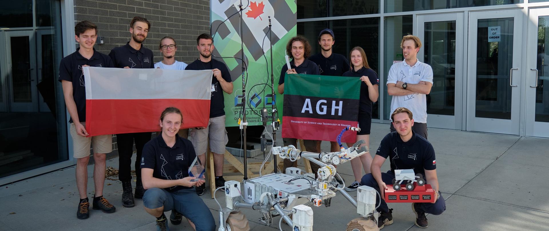 Przed wejściem do budynku stoi grupa młodych ludzi, trzymają polską flagą i zielono-czarno-czerwoną flagę z napisem ADH, przed nimi mały robot na czterokołowym podwoziu.