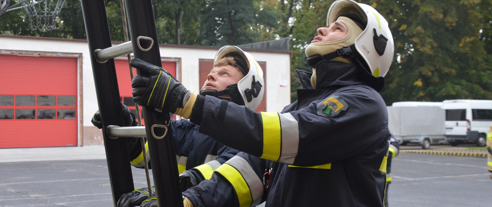 Szkolenie podstawowe strażaków-ratowników OSP. Plac komendy powiatowej w Rawiczu. Dwaj strażacy - kursanci sprawiają drabinę przystawną. Jedną rękę każdy z nich trzyma na bocznicy, a drugą - na szczeblu drabiny. Ćwiczący mają na sobie ubrania specjalne koloru ciemnego ze srebrno-żółtymi elementami odblaskowymi. Na ich głowach, zwróconych ku górze znajdują się hełmy koloru białego. W tle budynek, samochód z przyczepą oraz drzewa.