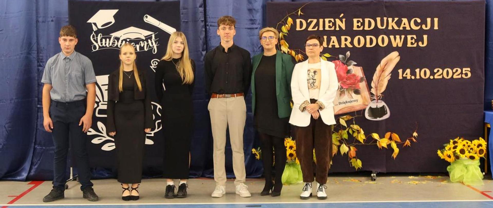 Czworo uczniów i dwie nauczycielki stoją na tle dekoracji z okazji Dnia Edukacji Narodowej.