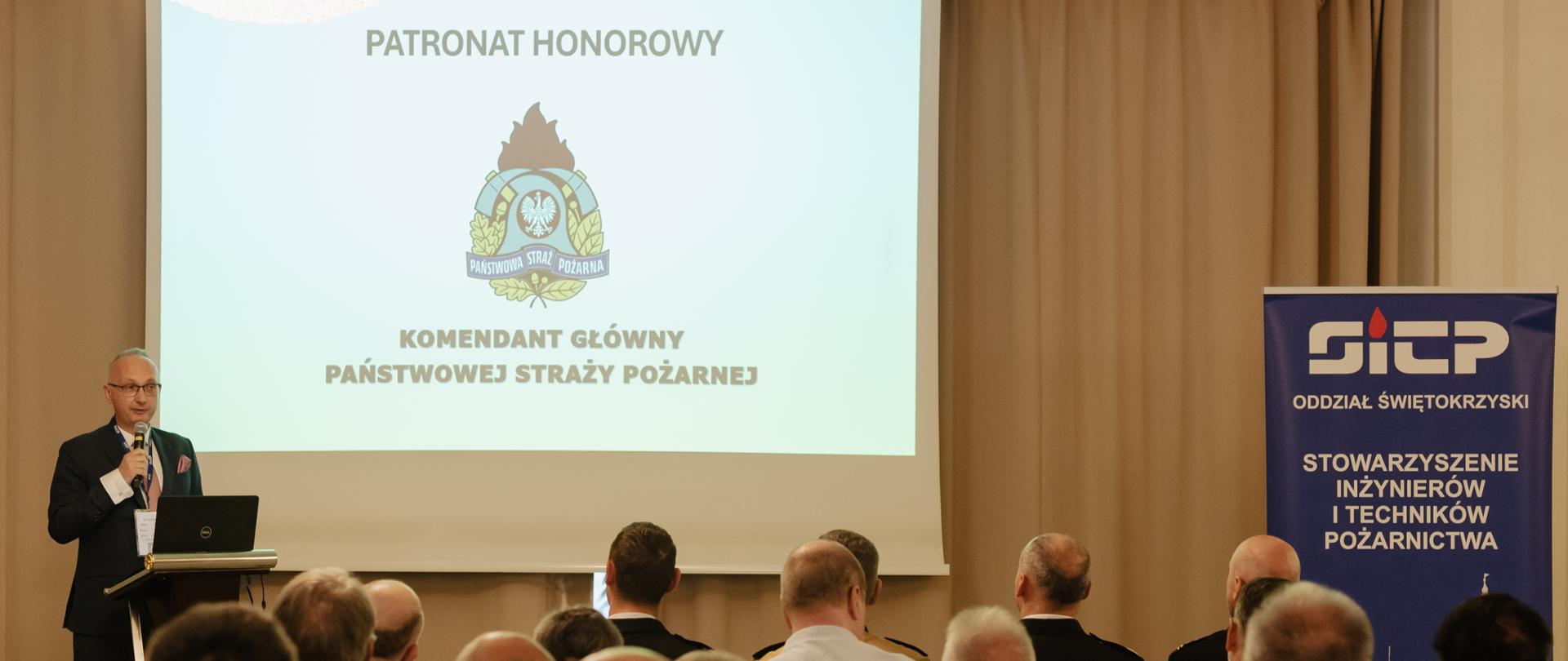 Na zdjęciu przemawia męzczyzna podczas konferencji, w tle na slajdzie logo PSP i napis patronat honorowy Komendnat Główny PSP