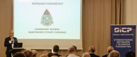Na zdjęciu przemawia męzczyzna podczas konferencji, w tle na slajdzie logo PSP i napis patronat honorowy Komendnat Główny PSP