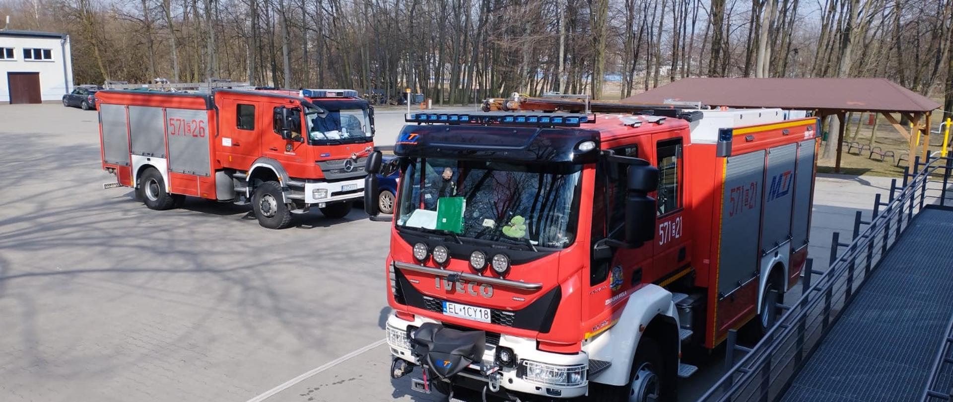 Zdjęcie przedstawia dwa samochody pożarnicze ustawione przed budynkiem. Z jednego samochodu rozwinięta jest linia gaśnicza. W tle widać drzewa. 