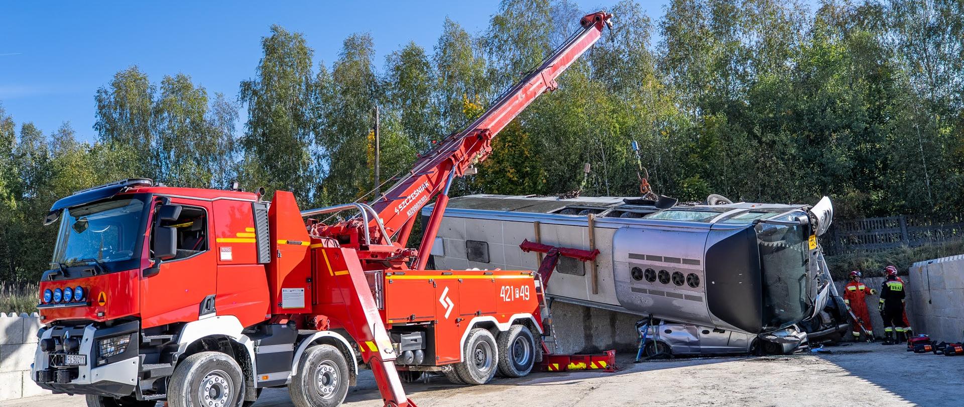 Czerwony ciężki samochód ratownictwa technicznego Państwowej Straży Pożarnej (z żurawiem hydraulicznym i numerem operacyjnym 421#49) stoi na placu manewrowym, stabilizowany za pomocą podpór. Pojazd podnosi za pomocą ramienia przewrócony na bok, uszkodzony srebrny autobus rejsowy, pod którym znajduje się zmiażdżony samochód osobowy. W tle widoczni są dwaj strażacy w pełnym umundurowaniu ochronnym oraz ściana zielonych drzew pod błękitnym niebem