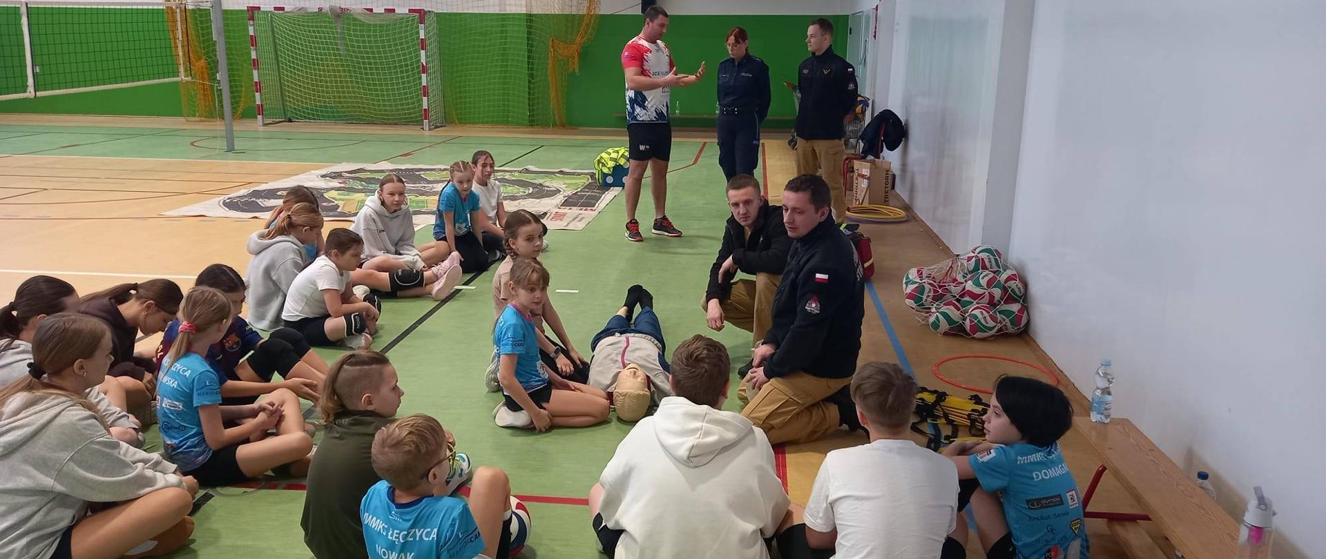 Grupa dzieci siedzi na podłodze w półkolu i uważnie obserwuje pokaz pierwszej pomocy. Dwóch strażaków, klęczy przy osobie leżącej na plecach (fantom) i demonstruje czynności ratownicze. Z boku stoją dwie osoby w mundurach służbowych oraz mężczyzna, którzy obserwują zajęcia. 