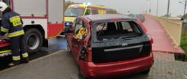 Na zdjęciu widać tył oraz lewy bok rozbitego pojazdu osobowego. Za rozbitym autem znajduje się samochód pożarniczy przy którym stoi strażak. W tle widać karetkę pogotowia oraz wiadukt drogowy