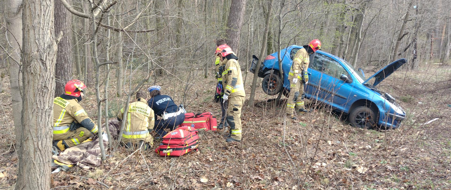 Zdjęcie przestawia zdarzenie drogowe w którym pojazd osobowy wypadł z drogi i znajduje się w lesie. Po prawej stronie zdjęcia znajduje się uszkodzony pojazd osobowy marki Peugeot 206 w kolorze niebieskim przy którym znajduje się jeden strażak. Po lewej stronie zdjęcia znajduje się osoba poszkodowana w pozycji leżącej, której udzielana jest kwalifikowana pierwsza pomoc przez czterech strażaków. Przy poszkodowanym znajduje się również policjant. 
