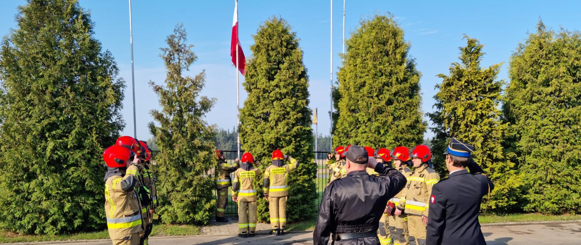Zdjęcie przedstawia strażaków Państwowej Straży Pożarnej w umundurowaniach specjalnych. Poczet flagowy składający się z trzech strażaków wciąga flagę na maszt. Odwróceni tyłem dowódca jednostki ratowniczo gaśniczej w umundurowaniu dowódczo sztabowym oraz komendant powiatowy w umundurowaniu wyjściowym salutują.