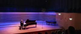 Mężczyzna stoi pryz fortepianie i mówi do mikrofonu, który trzyma w ręce. Z przodu widoczna od tyłu widownia z publicznością.
